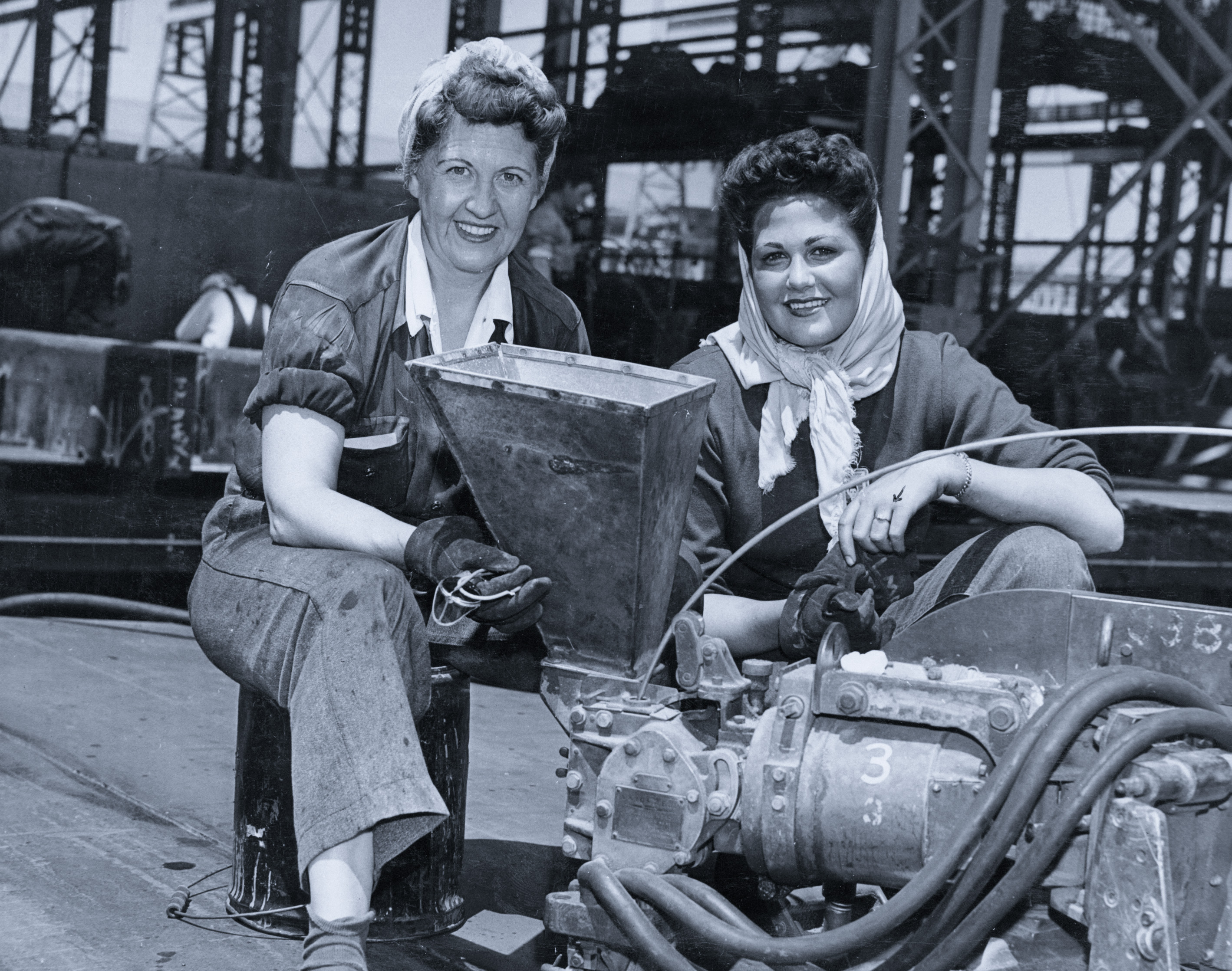 Historic photo of two women shipyard workers smiling at the camera. Industrial machines and equipment.