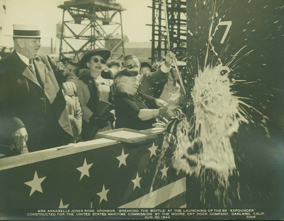 A group of men and women in dress clothes celebrate by breaking a champagne bottle across the ship.