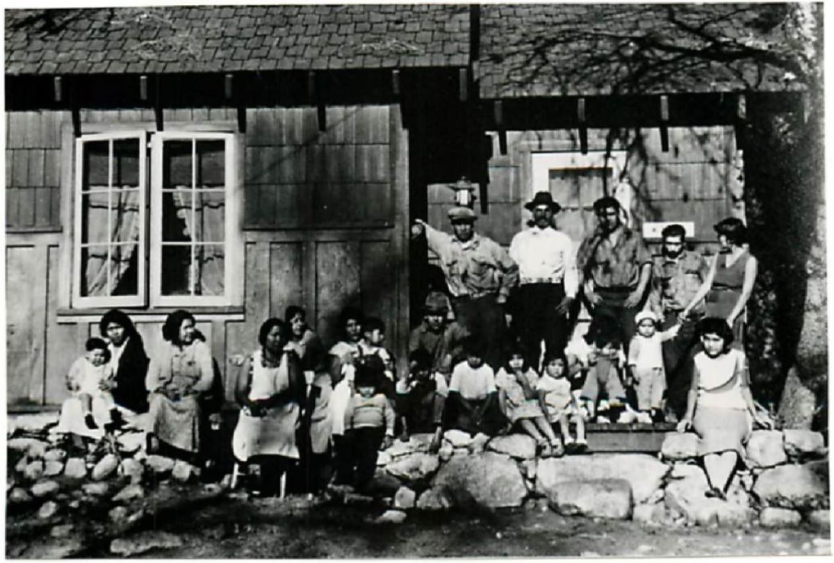 A black and white historical photo of a large group of people posing in front of a cabin in the New Village.