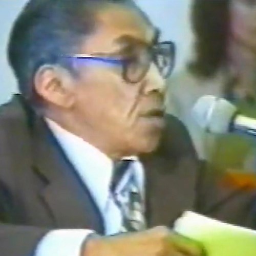 An Alaska Native man wearing thick glasses and a brown suit speaks into a microphone.