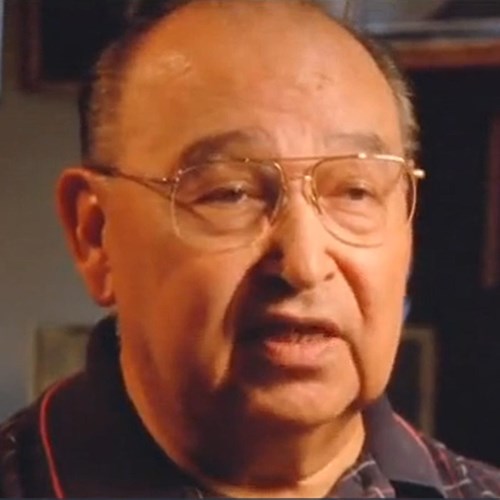 An Alaska Native man wearing a collared shirt and gold-rimmed glasses.