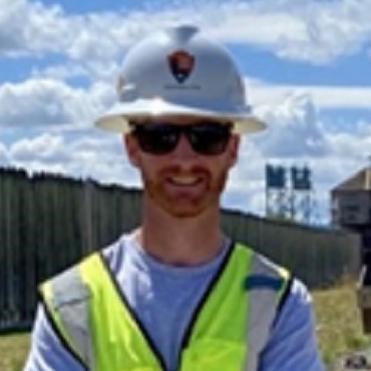 A white man smiles at the camera. He wears sunglasses, a white hard hat, and a safety vest.