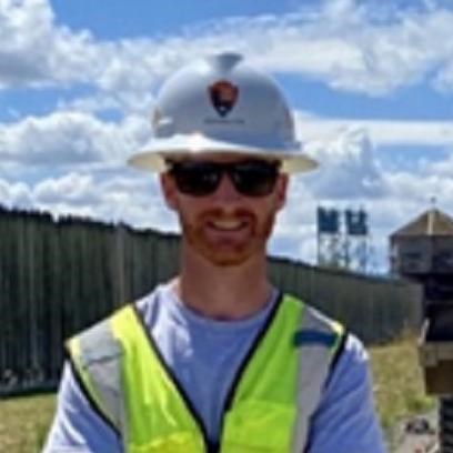 A white man smiles at the camera. He wears sunglasses, a white hard hat, and a safety vest.