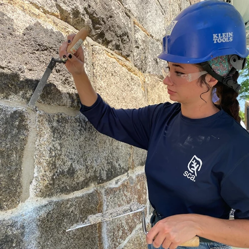 Person wearing a hardhat using a chisel against a granite wall's mortar