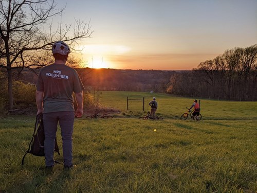 First State NHP volunteer, wearing a NPS volunteer shirt, stands in a field watching a sunset. Two mountain bikers stand at a distance in front watching the same sunset.