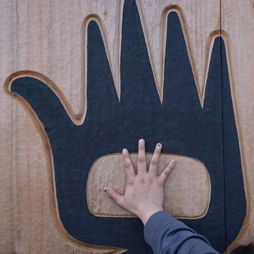 A hand placed over a carved wooden wall with a form line design of a hand.