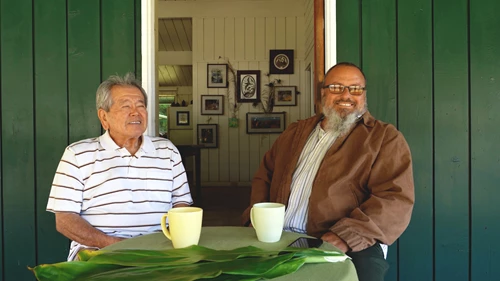 Two kūpuna (elders) sit at a table on a porch. The table is adorned with two coffee mugs