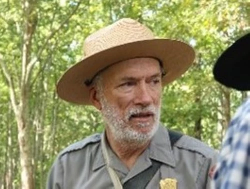 A ranger in a flat hat and a beard looks to his left.