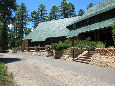 A rustic lodge with a broad green roof is surrounded by stone retaining walls and tall pines.