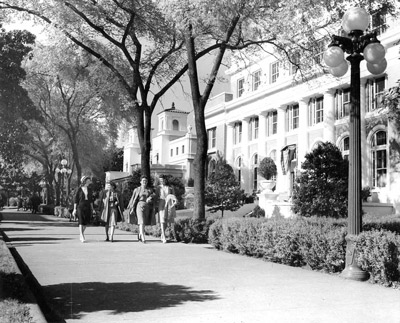 Four young women in 1940s-style skirts and jackets smile as they walk down a tree-lined walkway.