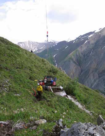man standing next to a pile of equipment dangling on a rope, on a grassy mountainside