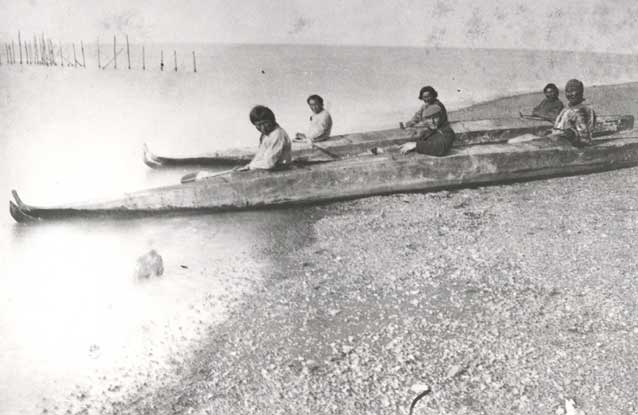 six people sitting in two long kayaks on a gravelly beach
