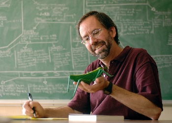 In front of a green chalkboard, a man makes handwritten notes from a scale model of a grasshopper