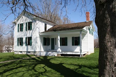A two story house with white siding, green shutters, and a porch on one side, surrounding by lawn.