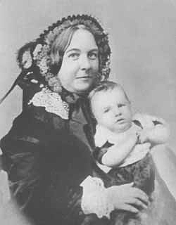 A women in a lace-collared dress and hat holds a baby to face the photographer.