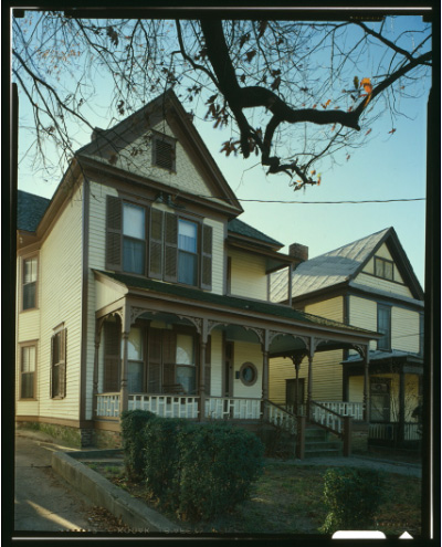 A narrow, two-story house with shutters, a porch, and low shrubs in front. 