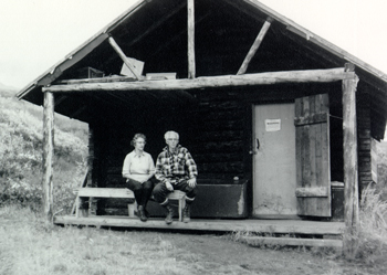 Adolph and his wife sit in front on a log cabin