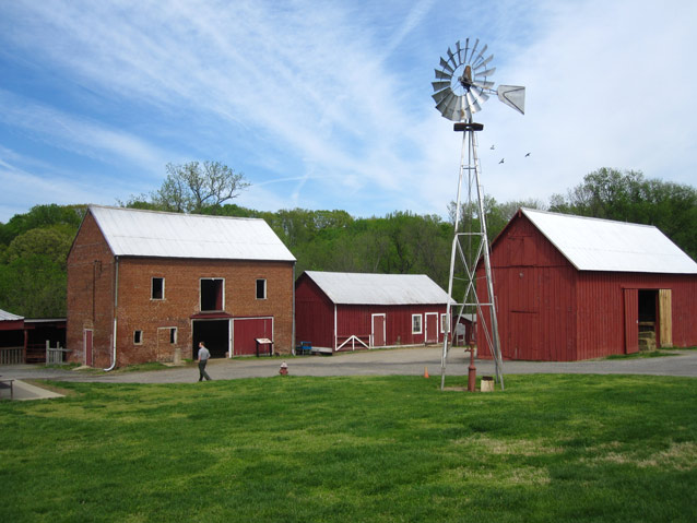 Wispy clouds above a windmill, farm buildings, and a bright green lawn. 