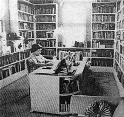 Black and white photo of a woman sitting in her desk surrounded by books 