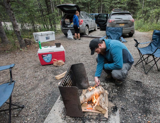 man tending a campfire while a young woman unpacks items from a nearby car
