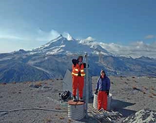 two women stand at the top of a seismic station with a snow-capped volcano in the distance