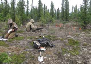 two people sit in a dirt clearing in a spruce forest