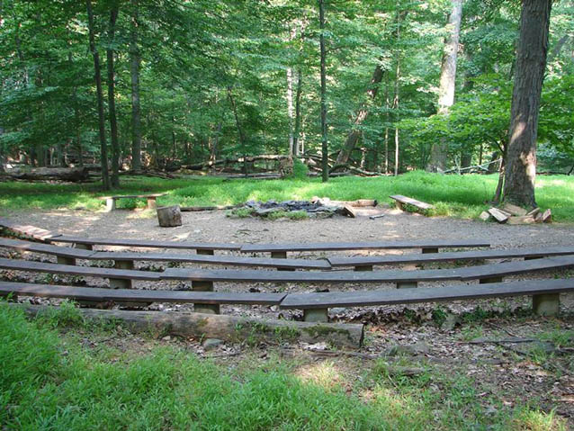 Wooden benches form a semi-circle around a campfire area, surrounded by forest