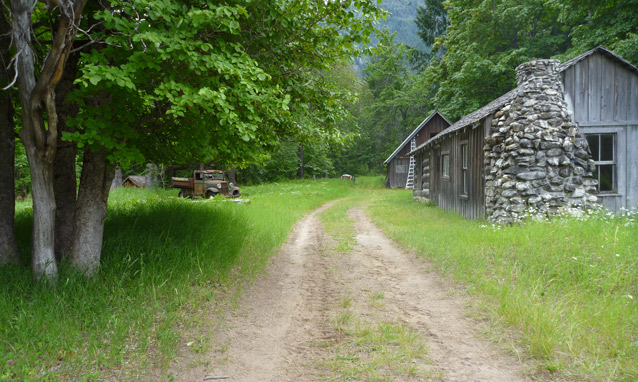 Tire tracks wear away grass in the driveway, leadidng to a wood and stone garage on the right