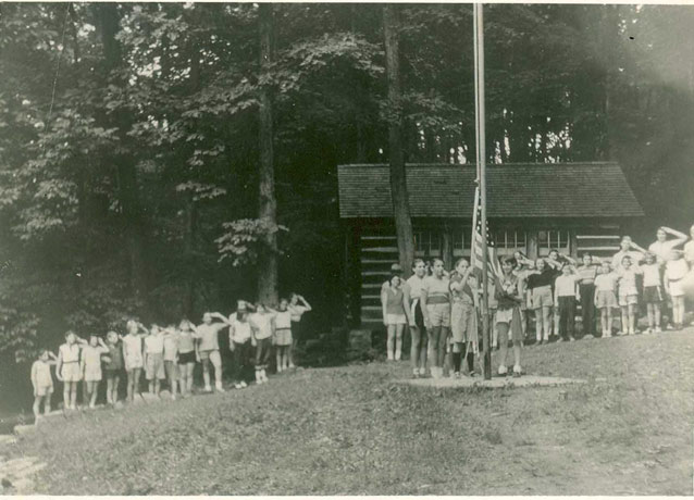 A crowd of young people, near a cabin, salutes as several girls attach the flag to a pole.