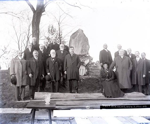 A group of men in overcoats and a seated woman in a long, dark dress pose beside a monument.