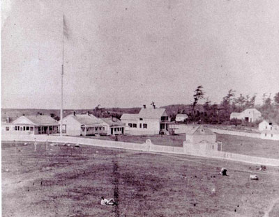 Grainy image of white buildings clustered behind a long picket fence on an open field