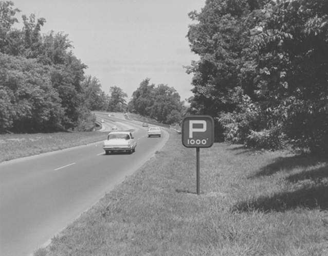 Historic image of a car driving on a parkway, bordered by a grassy shoulder and trees