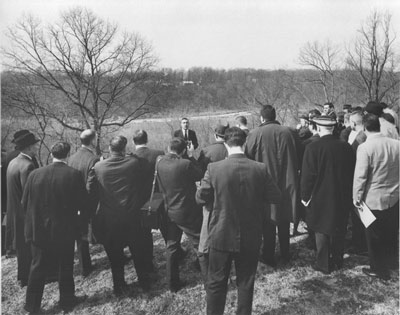 A man speaks to a crowd in suits and overcoats, all standing in front of a wide view.