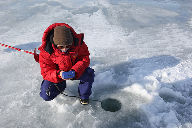 woman in red parka kneeling by a hole in ice