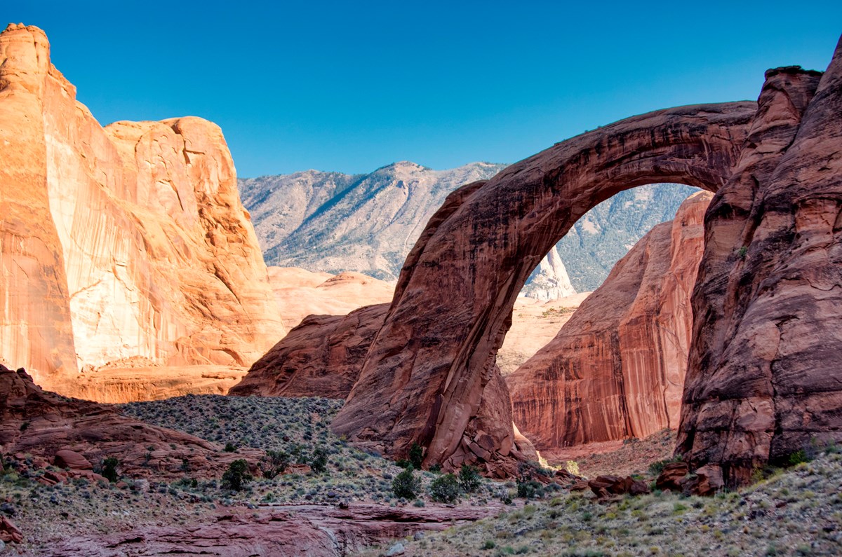 A natural stone arch sits between red rock canyon walls.  A mountain is seen through the arch.