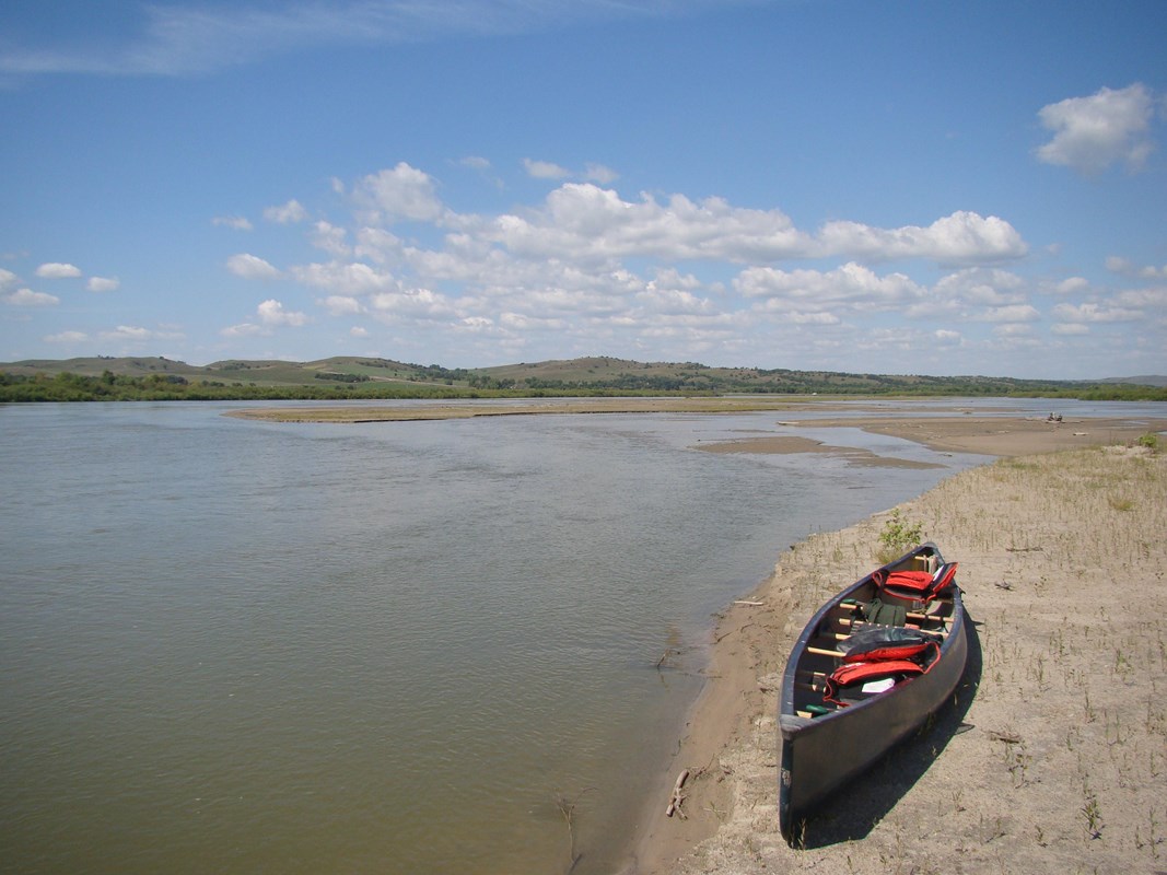 A canoe loaded with gear sits quietly on a sandy river bank with blue sky above.