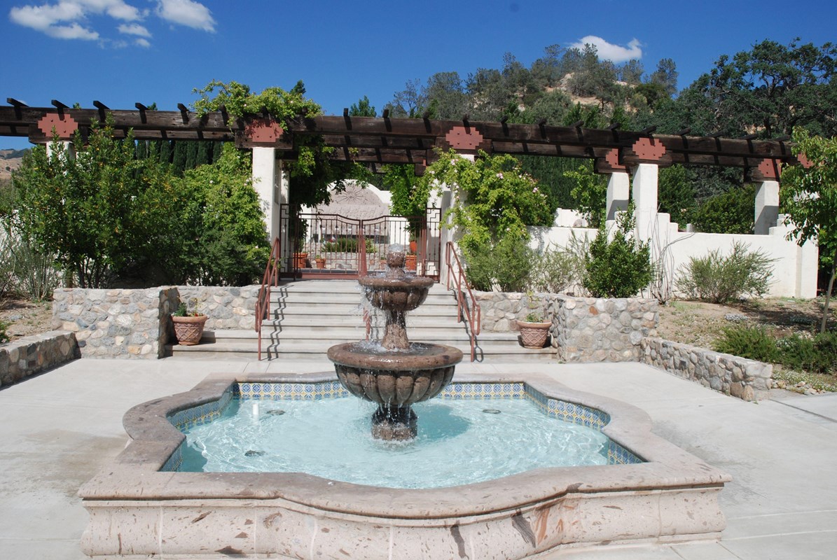 A two-tiered fountain in front of a set of stairs that go up to an arbor.