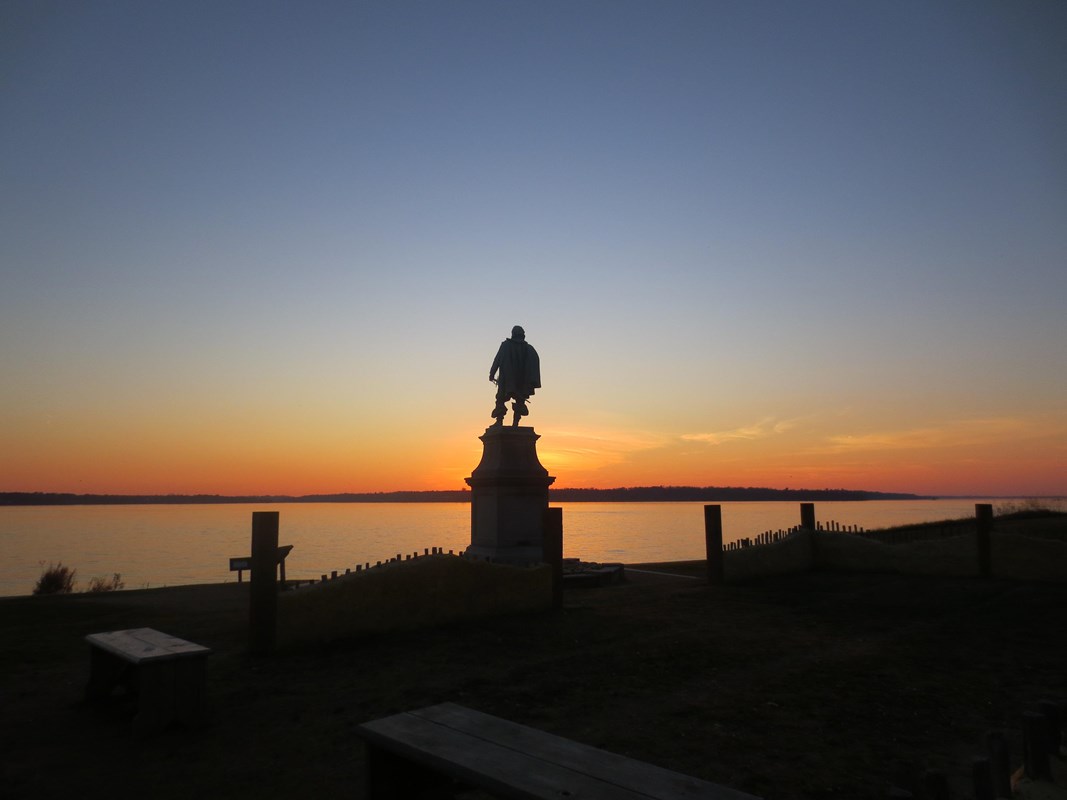 Statue on a pedestal silhouetted against a sunset over a river