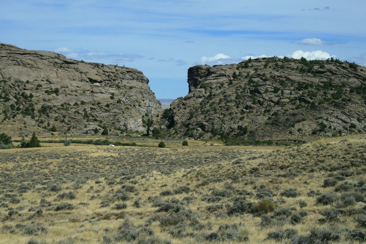 A tall rock buttress with a cleft in the center sits behind a sagebrush covered valley.