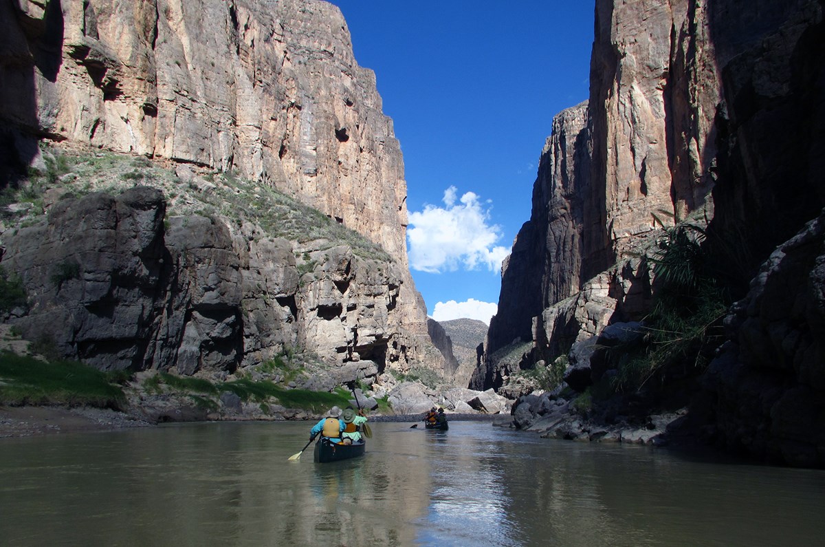 People in two canoes paddle on a river threading through high canyon walls. 