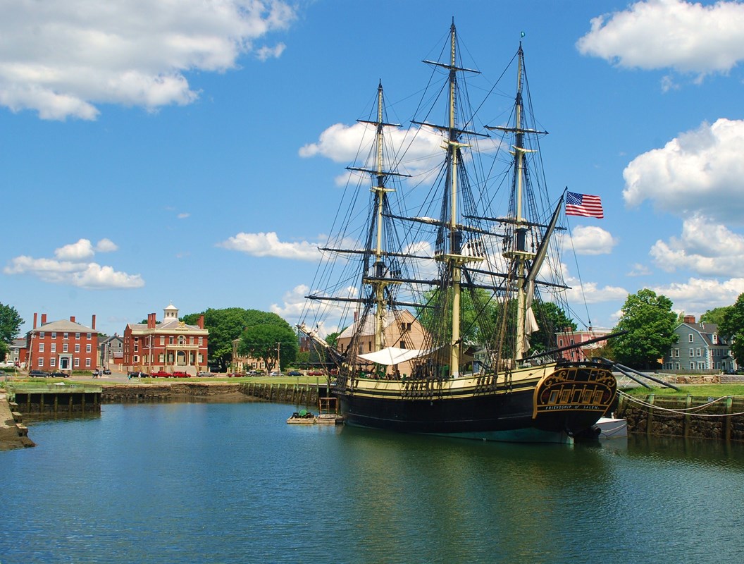A three mast tall ship on the water under a blue sky with red brick buildings on the shore.