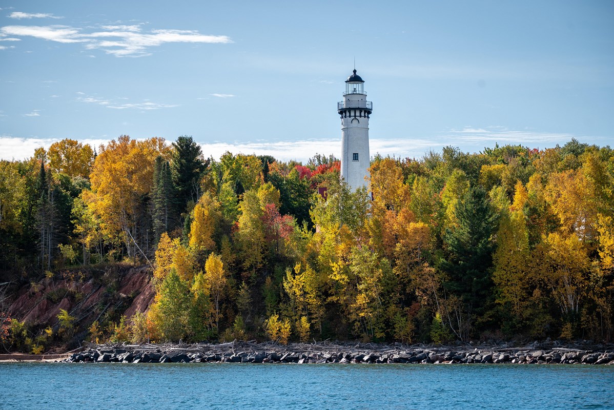 White lighthouse rising above colorful fall trees along a rocky shoreline under a blue sky.