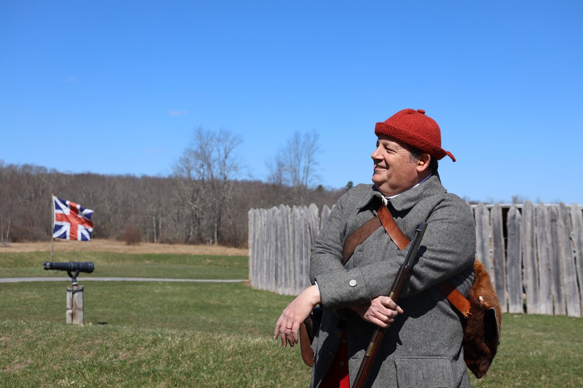 Man in 18th century flintlock musket stands in front of a round stockade fort with a British flag
