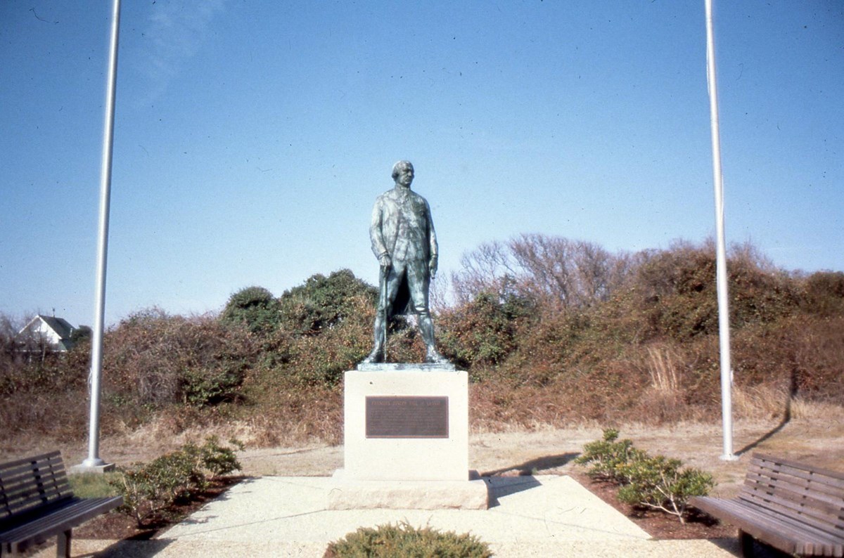 Patinaed statue of a military officer flanked by flagpoles and benches