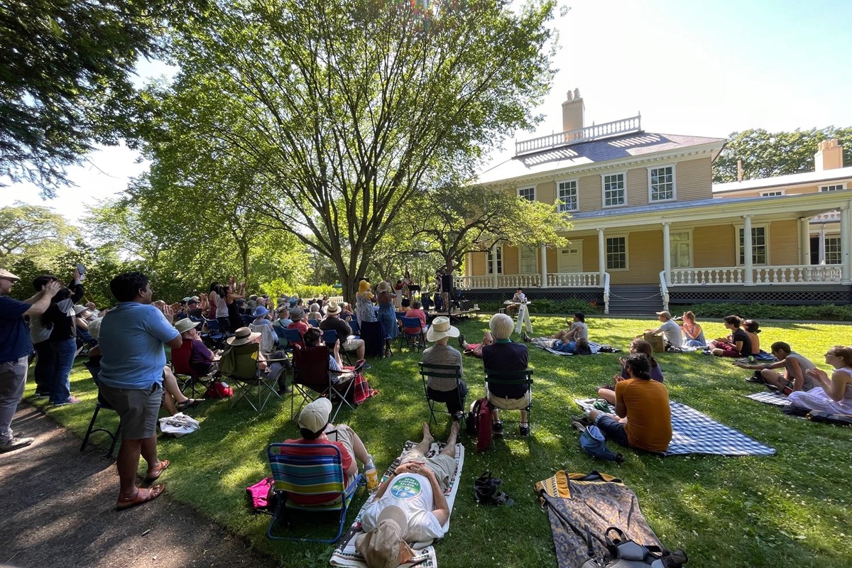 Crowd seated on a shaded lawn listening to an outdoor concert with yellow historic mansion