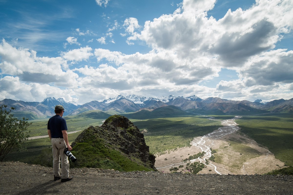 A man holding a camera looks out across a vast valley toward snow-capped mountains in the distance.
