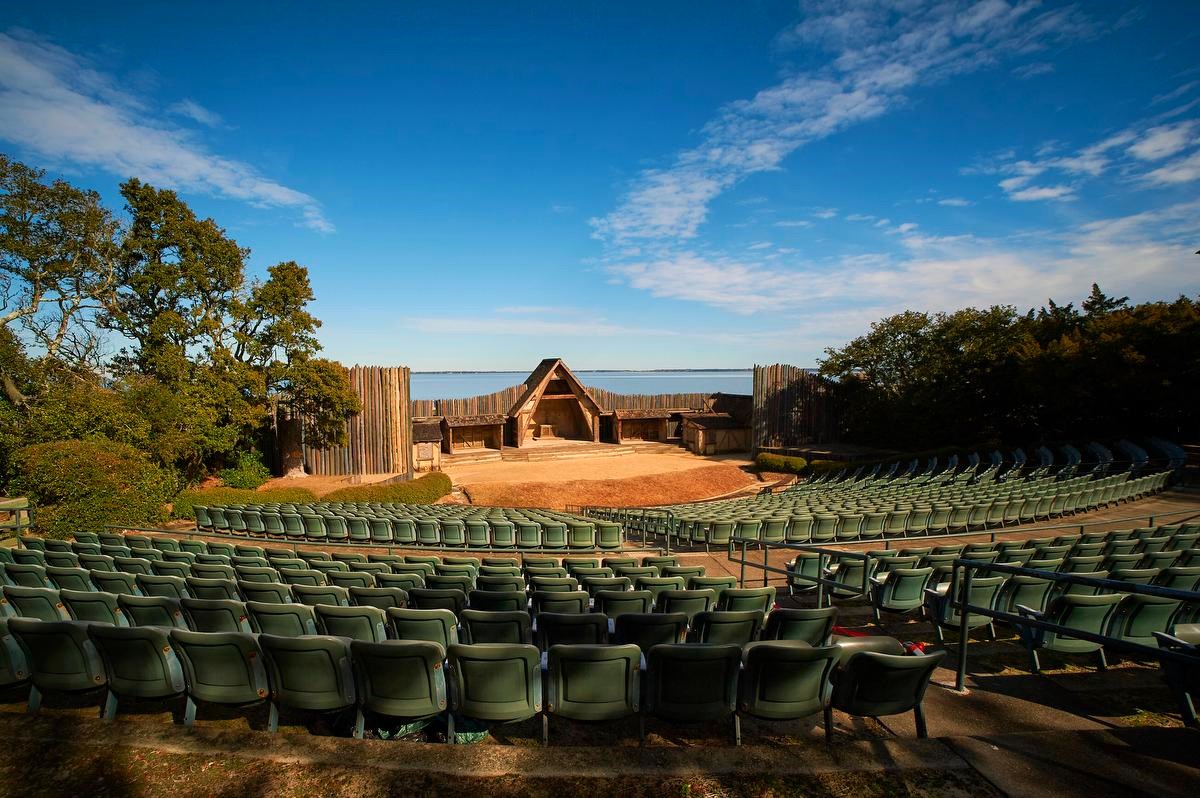 An outdoor theater on the shoreline with green stadium seats, sand stage, and wooden facade