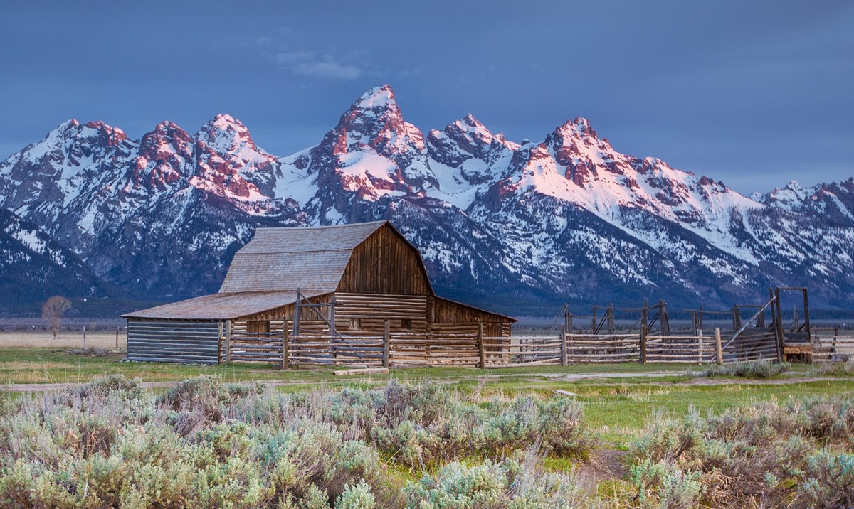 A wooden barn with snowy peaks in the background and green vegetation in the foreground