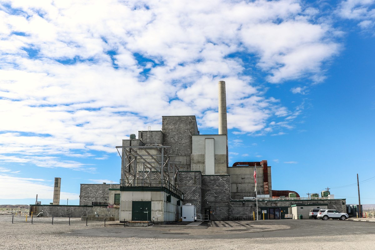 Gray factory building like stacked boxes with single taller smokestack beneath cloudy blue skies. 