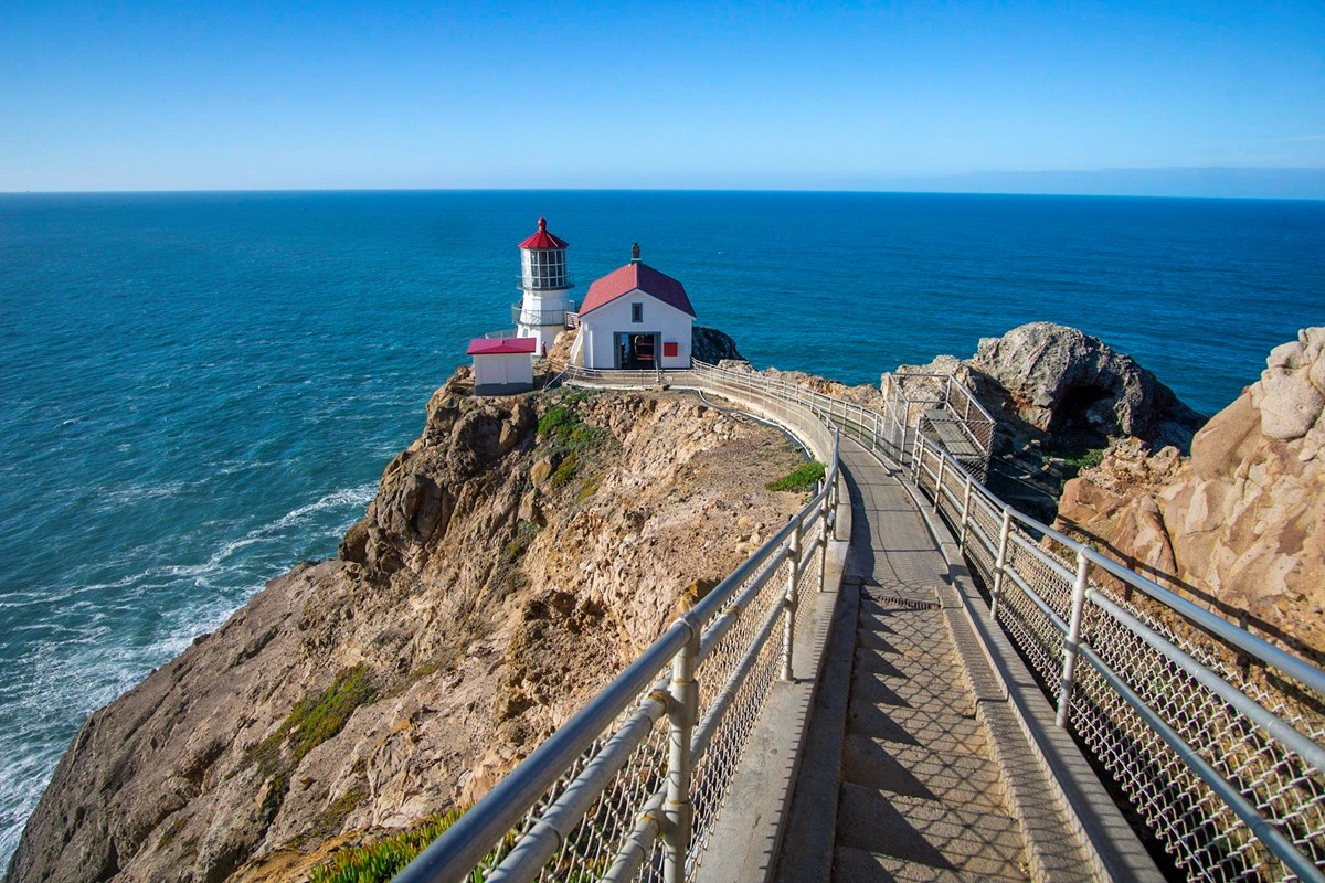 A long concrete staircase down to a red roofed, white lighthouse on cliffs above the ocean.